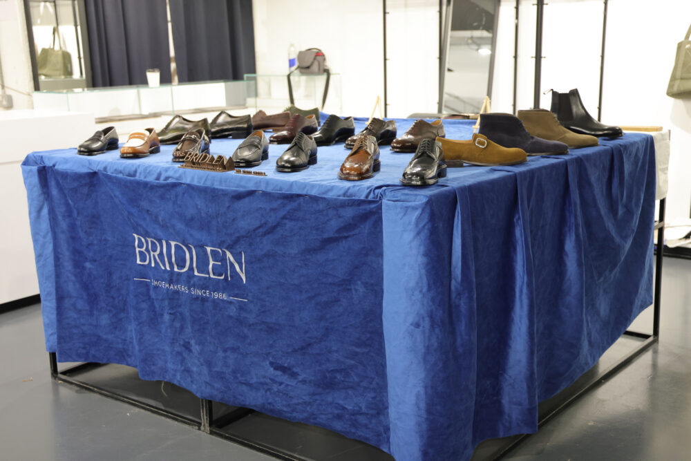 A table covered with a blue cloth displays various styles of shoes, including leather and suede options, with the brand name "Bridlen" prominently featured.