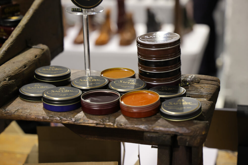 A wooden shelf displays various round tins of Saphir shoe polish in colors including black, brown, orange, and purple.