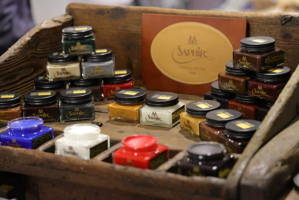 A wooden display holds several jars of Saphir shoe cream in various colors, including blue, red, and green.