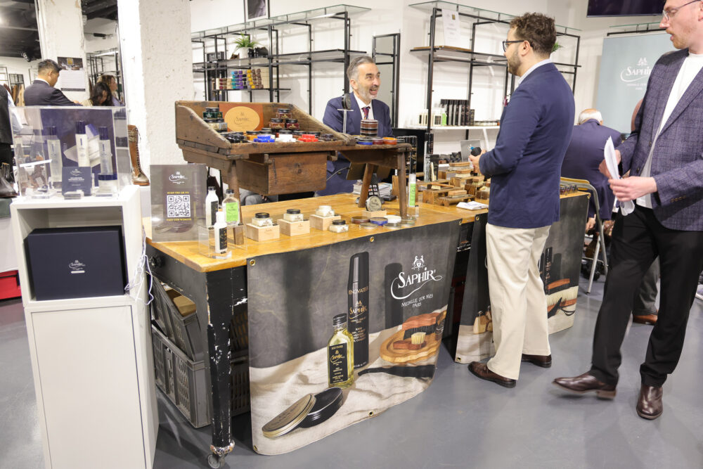 A vendor displays Saphir leather care products on a wooden table with various jars and bottles, while a customer engages with a representative.