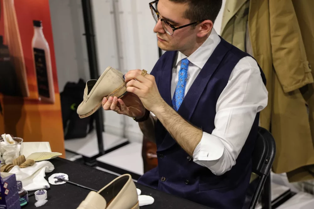 A man in a navy vest and white shirt is cleaning a beige shoe with a cloth while sitting at a table.