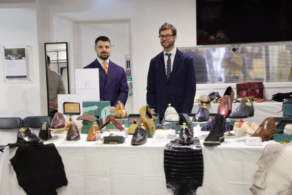 Two men in tailored suits stand behind a table displaying a variety of leather shoes in different colors and styles.