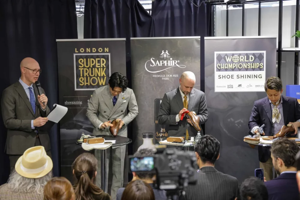 Four men in formal attire are demonstrating shoe shining techniques on brown leather shoes during a shoe shining competition.