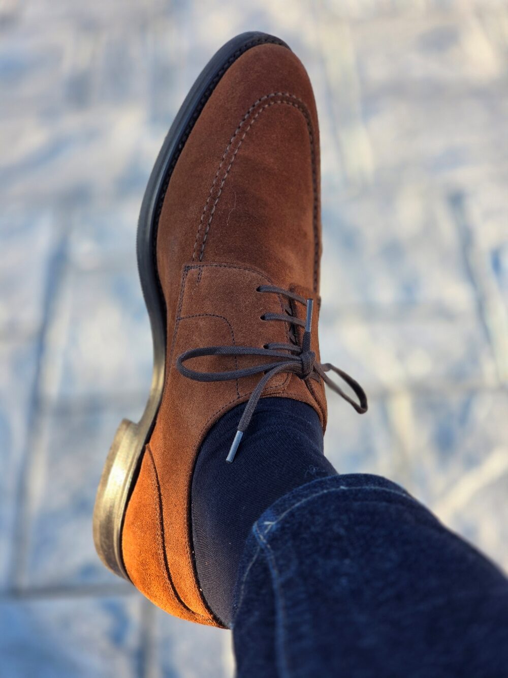 A close-up of a brown suede dress shoe with laces, worn with dark socks and denim jeans.