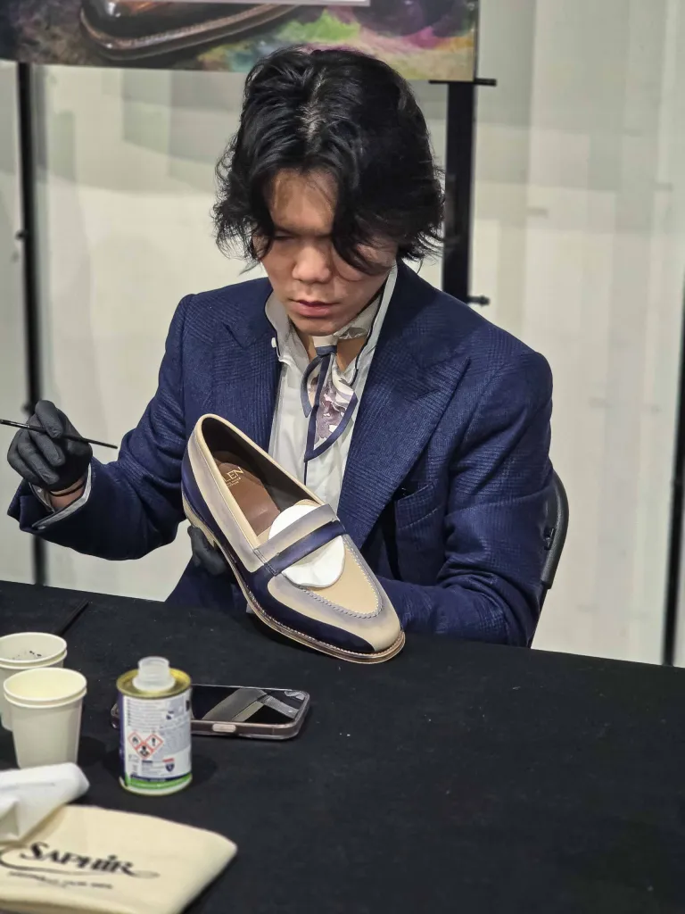 A person in a blue suit carefully paints a two-tone leather shoe, featuring beige and navy colors.