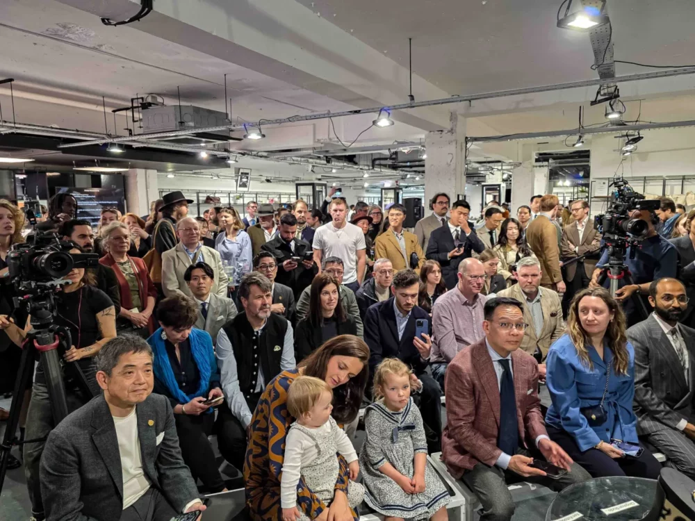 A diverse audience seated in formal attire, including a man in a gray suit and a woman holding a toddler, at an event with cameras present.