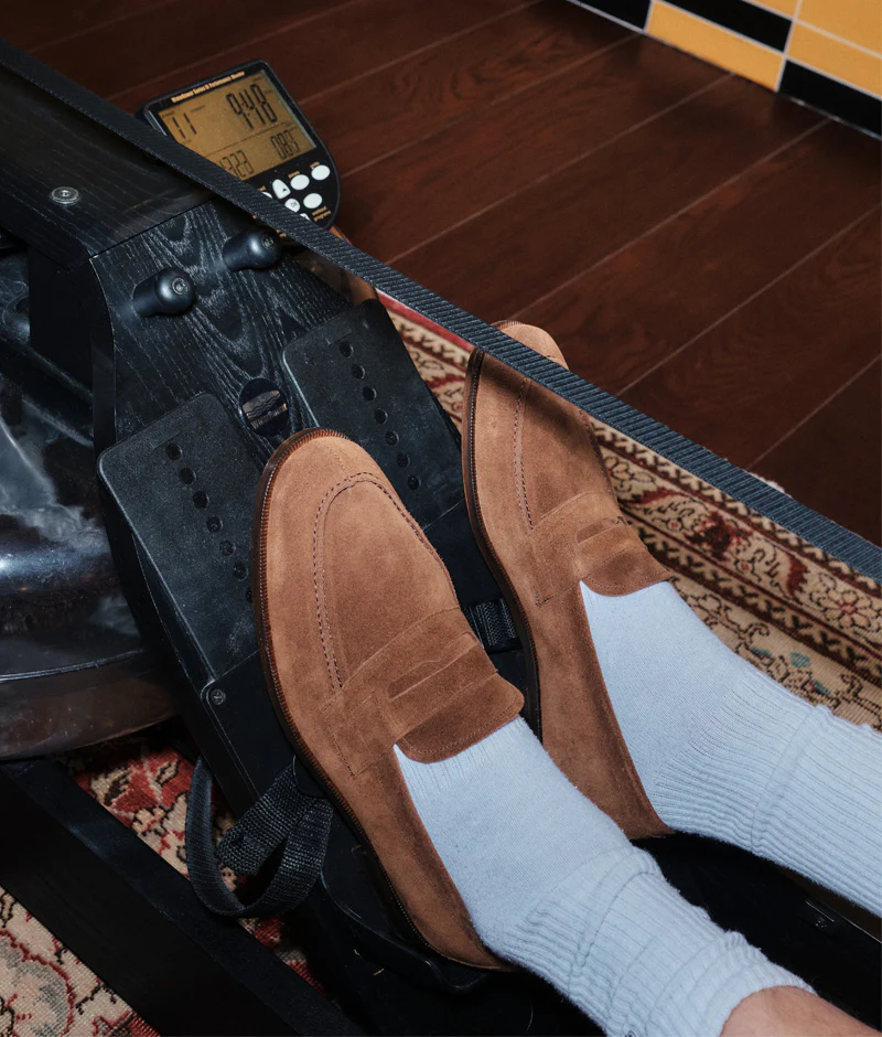 A pair of feet wearing brown suede loafers and white socks rests on the footplates of a rowing machine.
