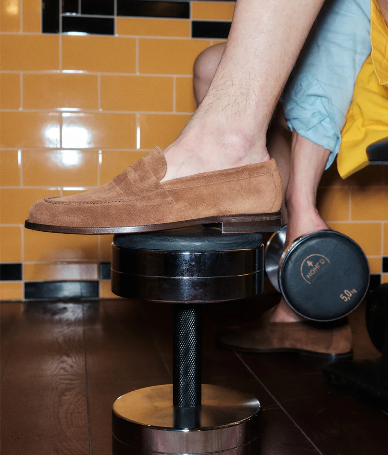 A foot wearing a brown suede loafer rests on a black weightlifting plate.