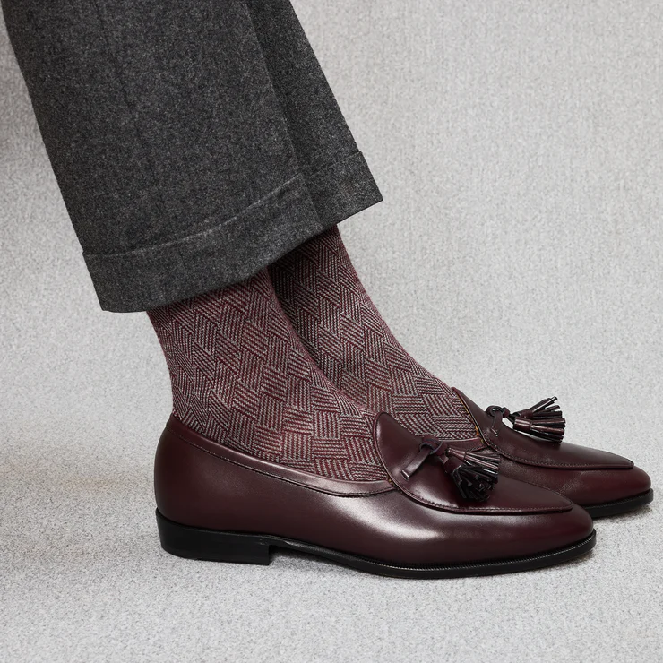 A close-up of a pair of burgundy leather loafers with tassels, worn with patterned maroon socks and gray trousers.