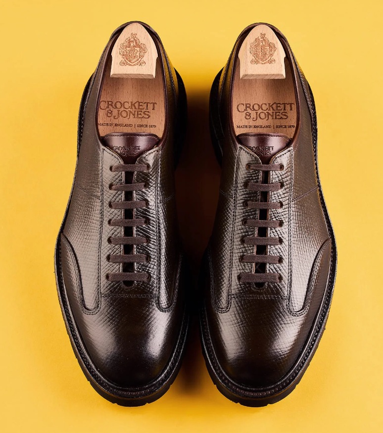 A pair of dark brown leather shoes with a textured finish and laces, featuring a wooden insole marked with "Crockett & Jones."