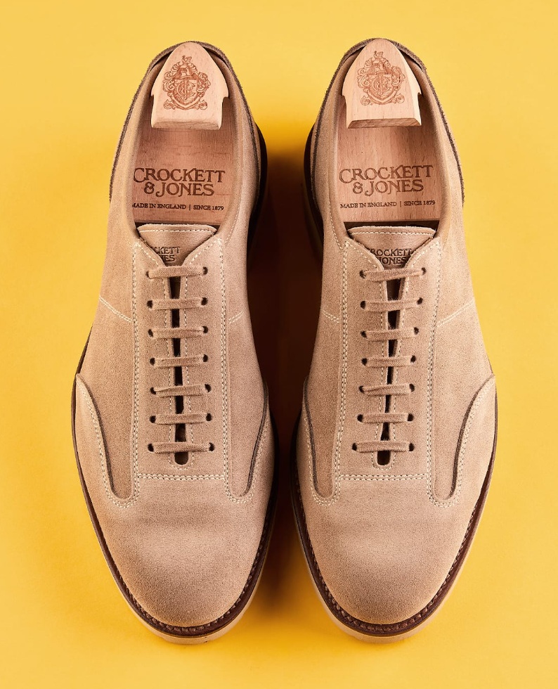 A pair of beige suede shoes with a lace-up design and wooden shoe trees labeled "Crockett & Jones."