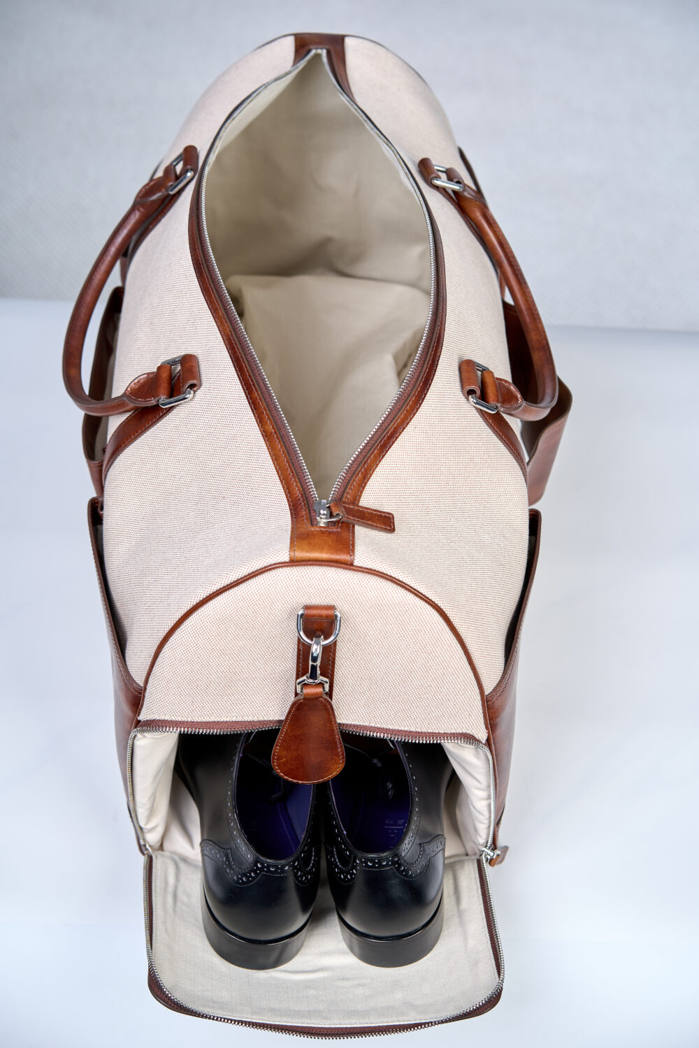 A beige duffel bag with brown leather accents, featuring a compartment holding a pair of black dress shoes.