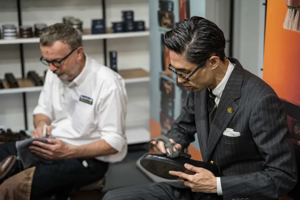Two men are polishing shoes, one in a white shirt and glasses, and the other in a black pinstripe suit with glasses.