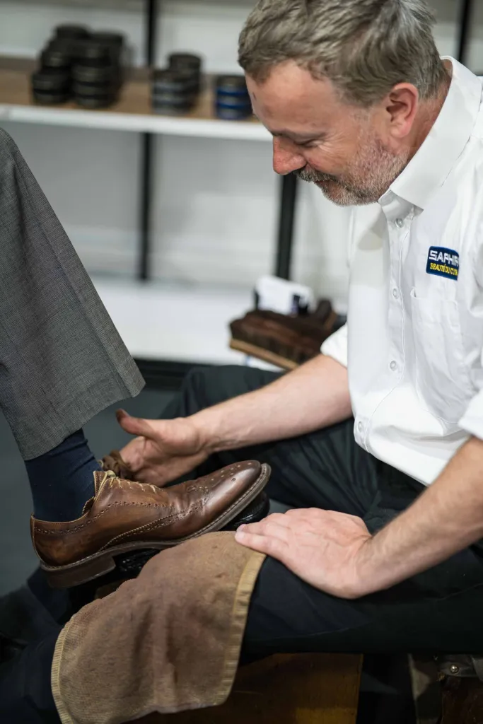 A shoeshiner polishes a brown leather shoe while seated, wearing a white shirt.
