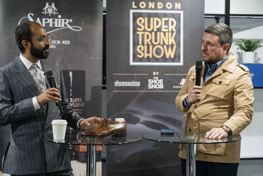 Two men are speaking at a presentation, with one holding a brown dress shoe and a coffee cup on a glass table.