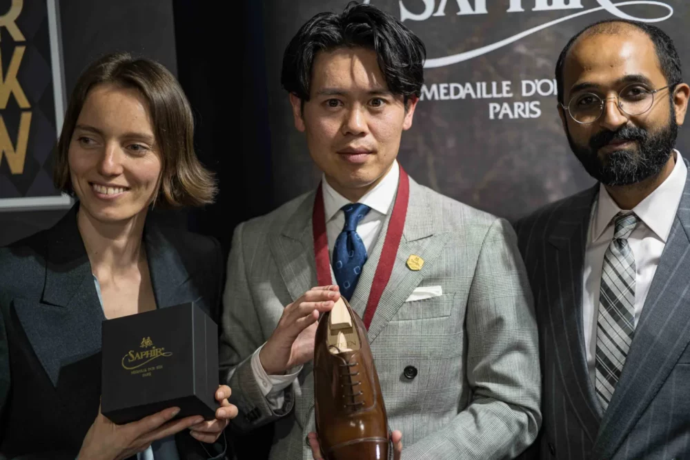 Three people stand together, with the central figure holding a brown leather shoe and a medal, while one person holds a black Saphir box.