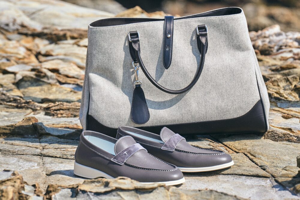 A gray handbag with dark leather accents is placed next to a pair of gray slip-on shoes.