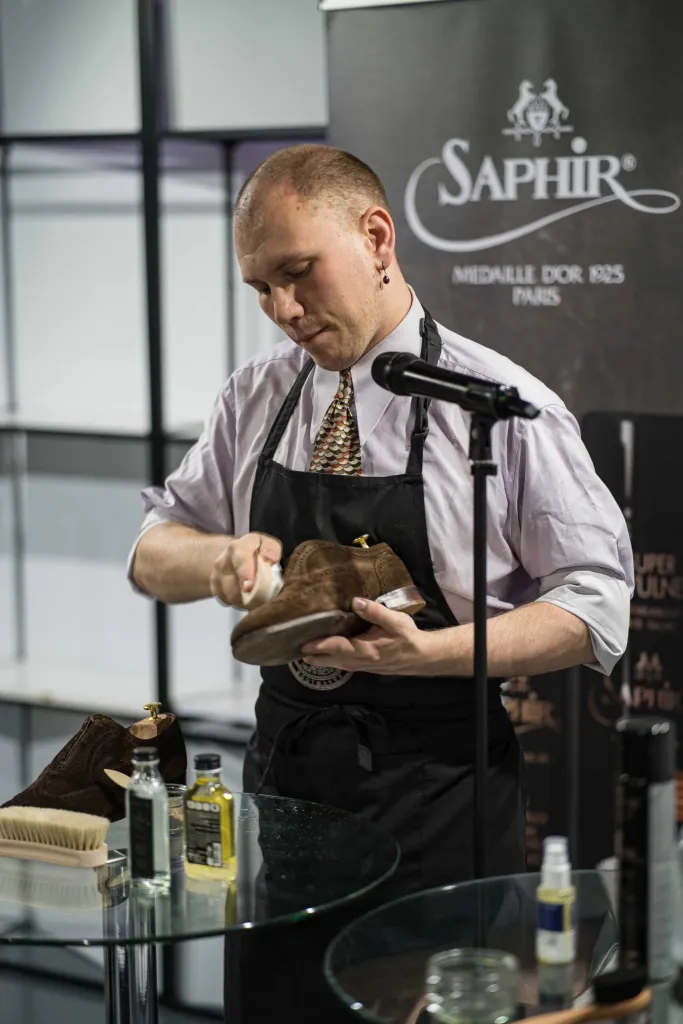 A person in an apron is polishing a brown suede shoe while holding a brush and surrounded by shoe care products.