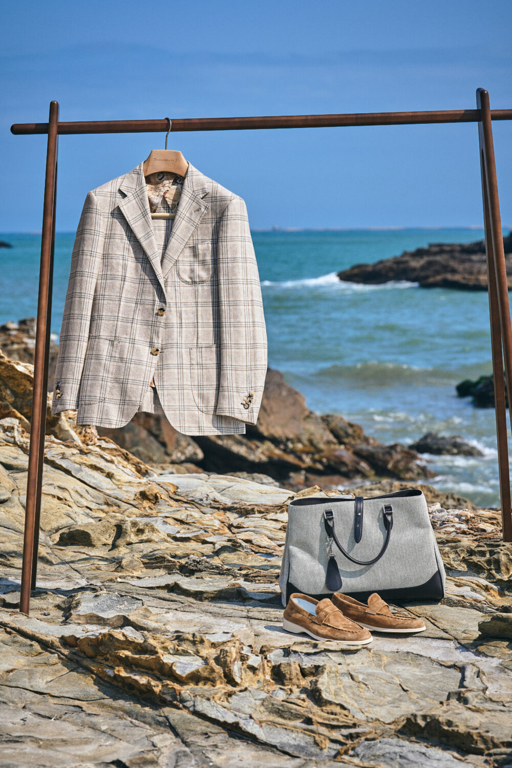 A beige plaid blazer is displayed on a wooden hanger alongside a gray bag and brown loafers on rocky terrain.
