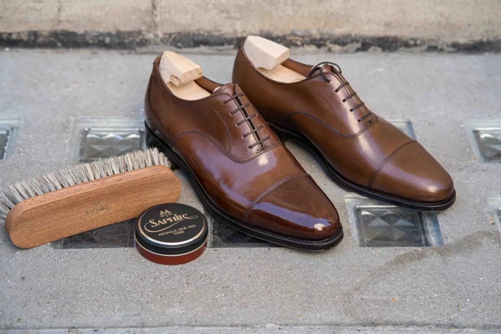 A pair of polished brown leather shoes with wooden shoe trees, accompanied by a wooden brush and a tin of shoe cream.