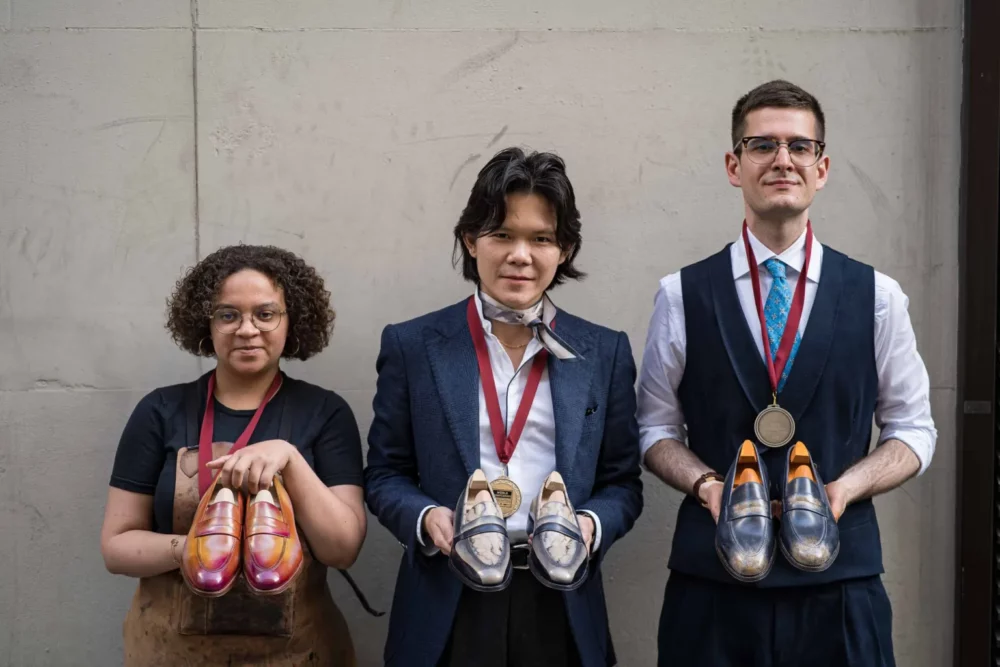 Three individuals hold handmade shoes: one with orange and pink shoes, another with gray and white shoes, and the third with dark blue shoes featuring orange insoles.