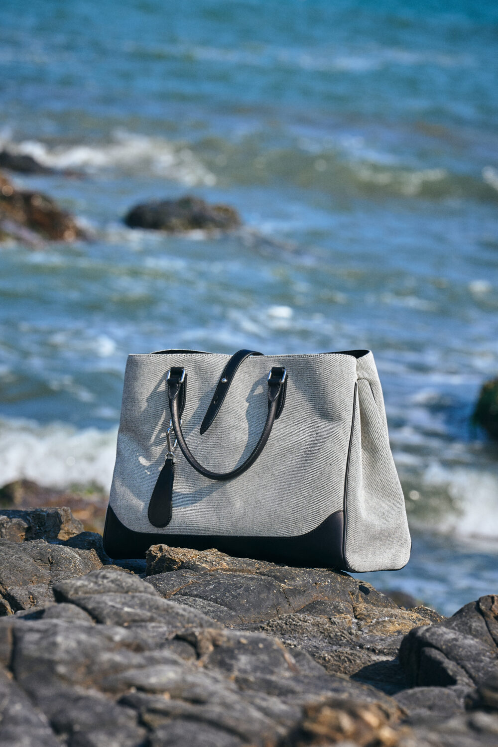 A gray and black tote bag with dual handles rests on rocks by the water.