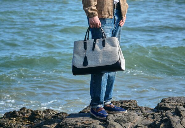 A person stands on rocks by the water, holding a gray and black handbag.