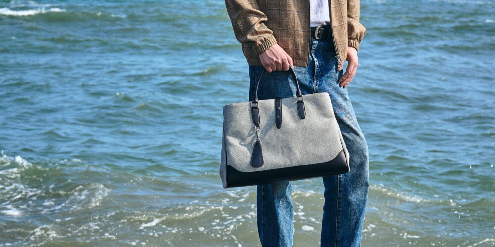 A person stands by the water holding a gray and black handbag.
