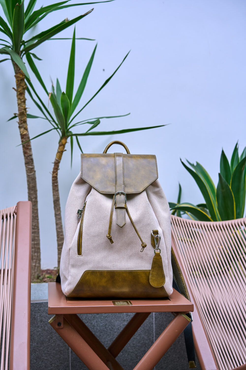 A beige backpack with a brown leather flap and details sits on a wooden stool.