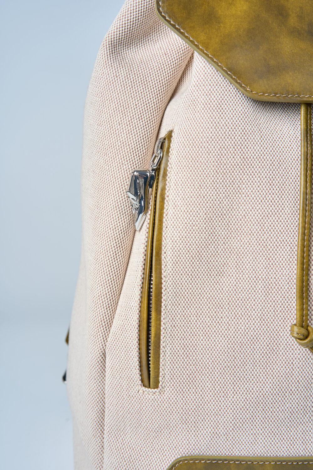 A close-up of a beige backpack featuring a brown leather flap and a silver zipper.