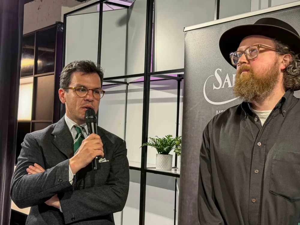 A man in a gray suit and glasses speaks into a microphone while another man with a beard and black shirt looks on.