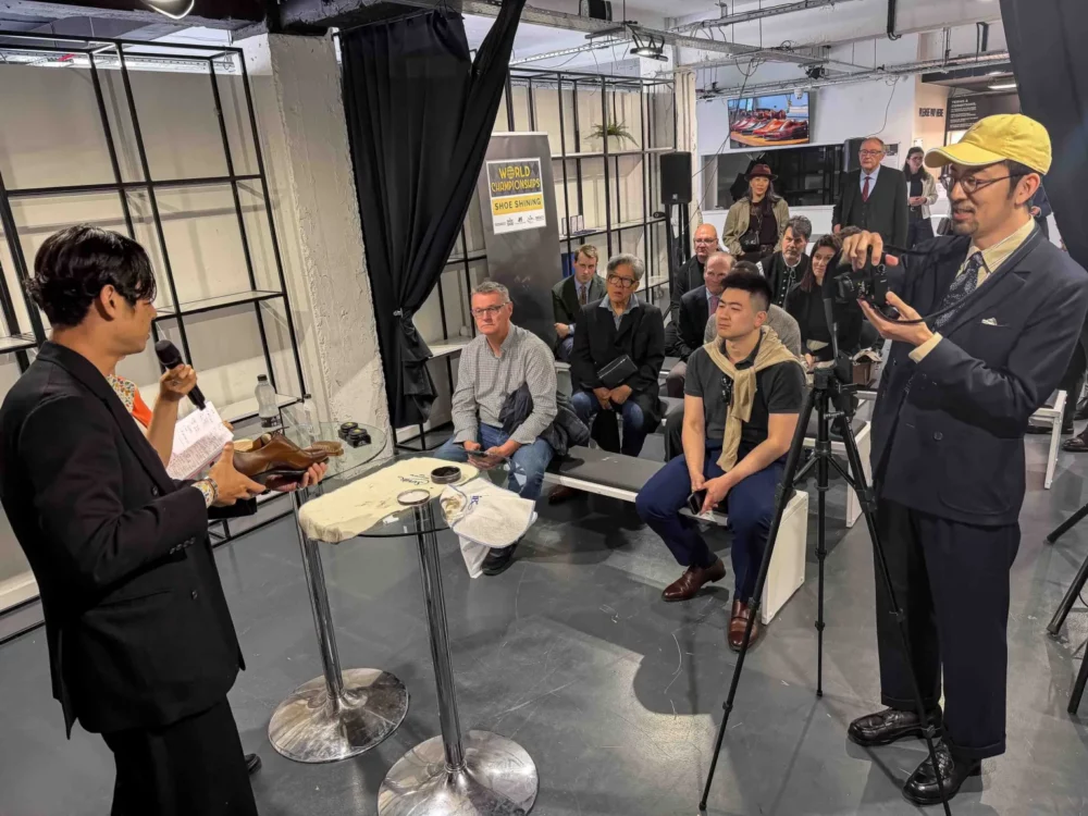 A man in a black suit holds a brown shoe and microphone while demonstrating shoe shining techniques to an audience.