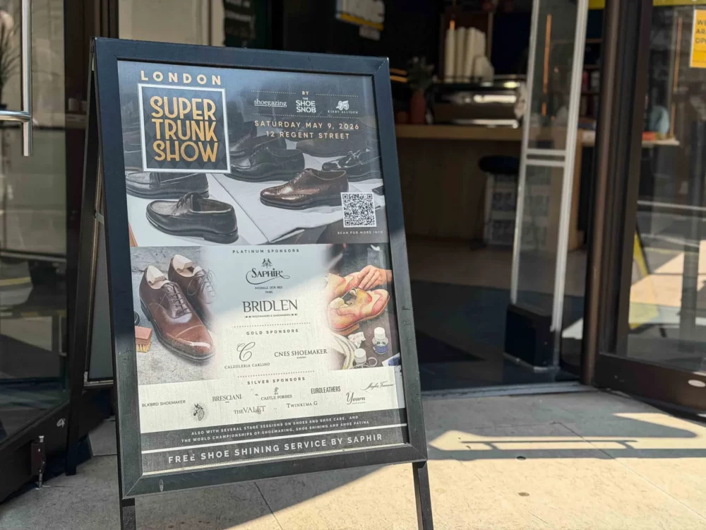 A black sandwich board sign advertising the London Super Trunk Show with images of shoes and details about sponsors and a free shoe shining service.