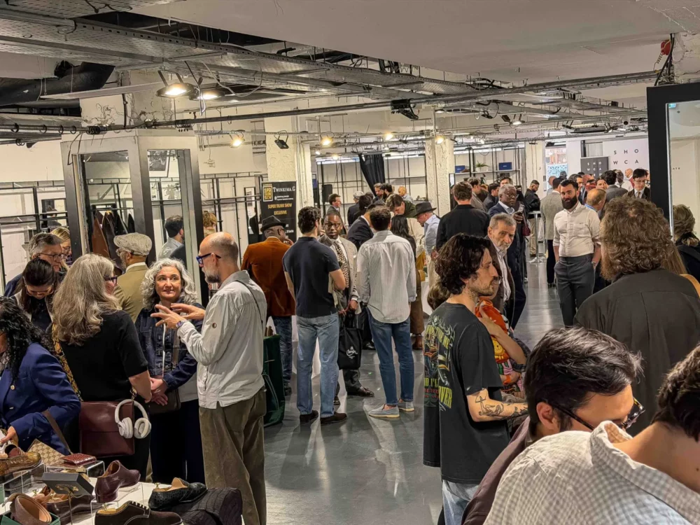 A diverse crowd of people engages in conversation at a footwear exhibition, with various styles of shoes displayed in the foreground.