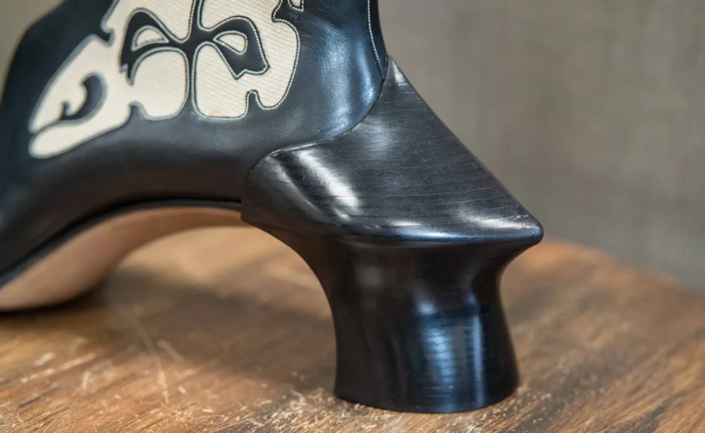 A close-up of a black high-heeled shoe featuring a unique sculpted heel and cream-colored decorative stitching.
