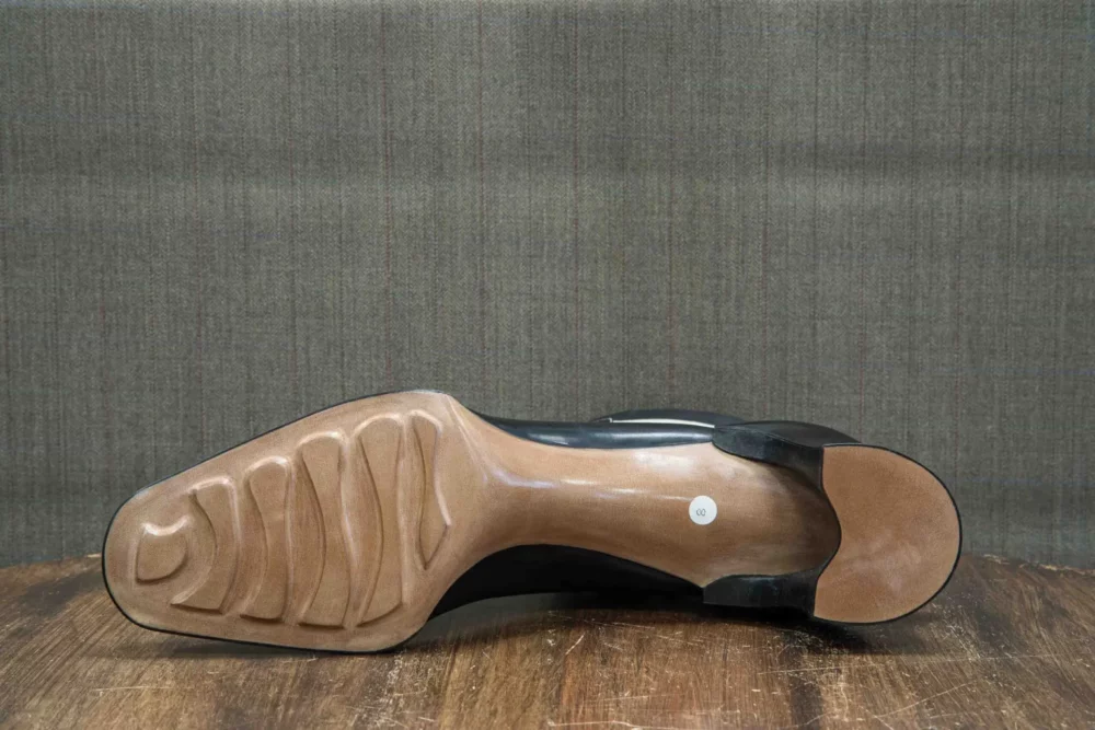The image shows the underside of a black shoe with a smooth tan sole and textured grooves for grip.
