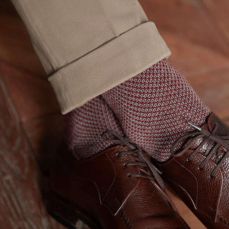 A close-up of a brown leather shoe paired with a red and gray patterned sock, above light beige pants.