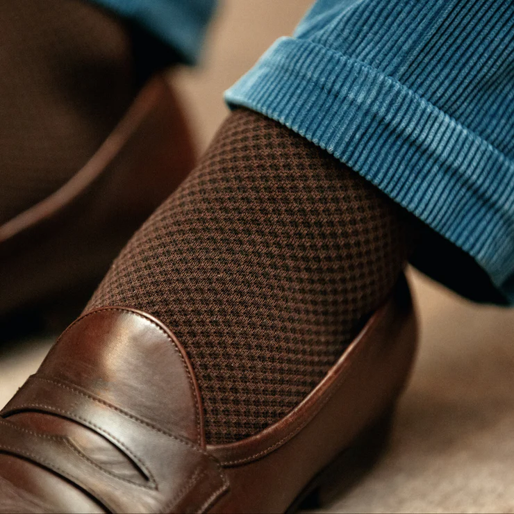 A close-up of a brown patterned sock paired with a brown leather loafer and blue corduroy pants.