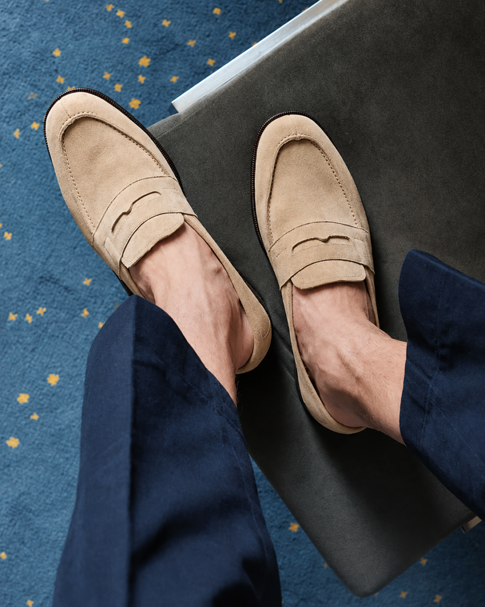 A pair of beige suede loafers worn with dark blue pants.