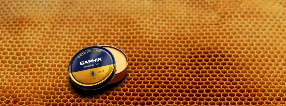 A round tin of Saphir leather cream in blue and gold sits on a honeycomb-patterned surface.