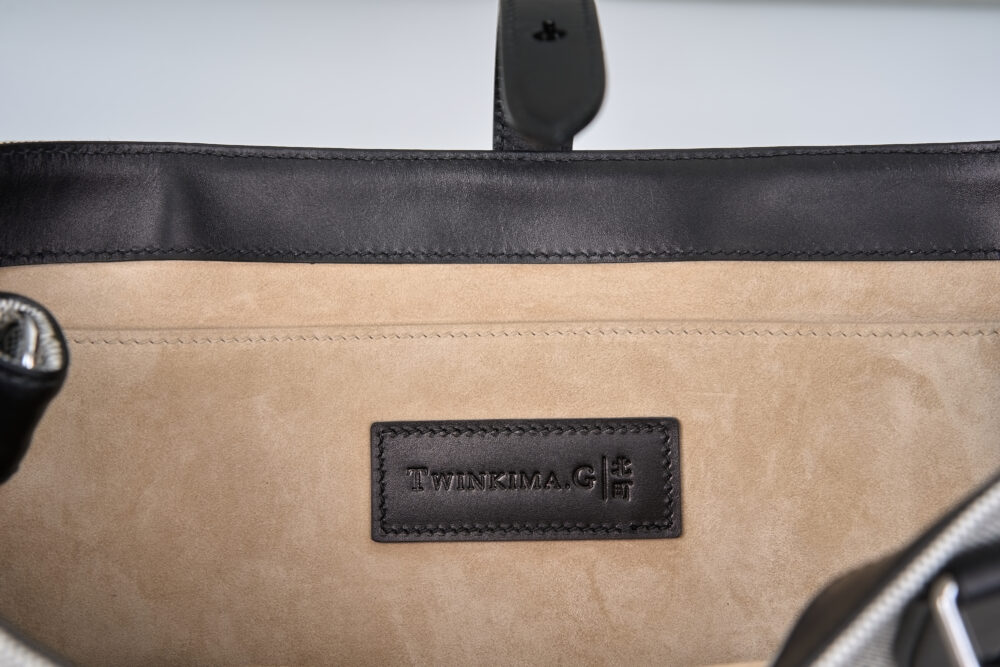 An interior view of a handbag featuring a black leather trim and a beige suede lining with a black embossed label.