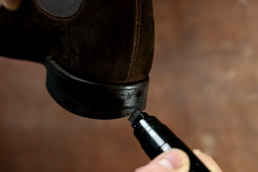 A hand holds a black marker being used to touch up the black heel of a brown suede shoe.