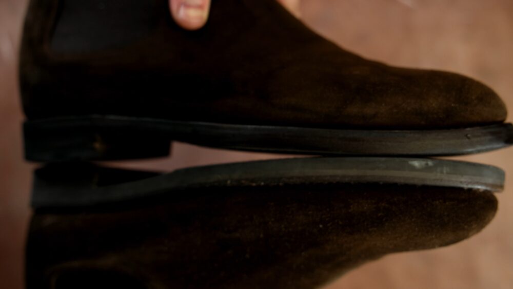 A pair of dark brown suede shoes with a black sole, shown from the side with a hand holding one.