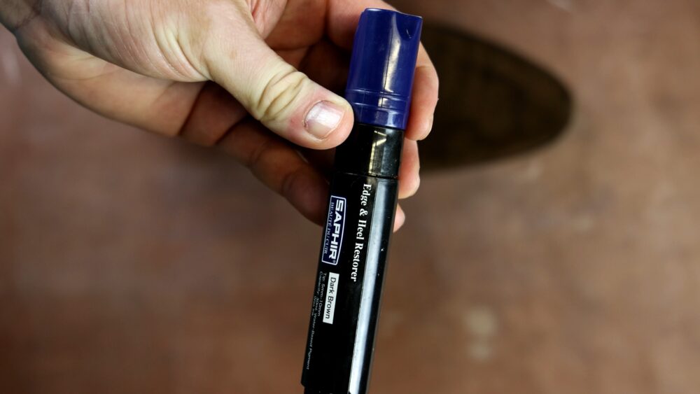 A hand holds a black and blue Saphir edge and heel restorer marker labeled "Dark Brown."