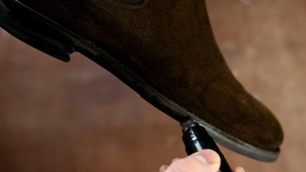 A person is applying a black shoe polish to a brown suede boot.