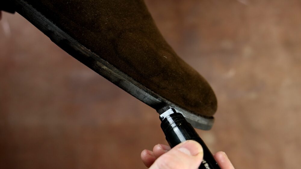 A person is applying a black shoe marker to the edge of a brown suede boot.
