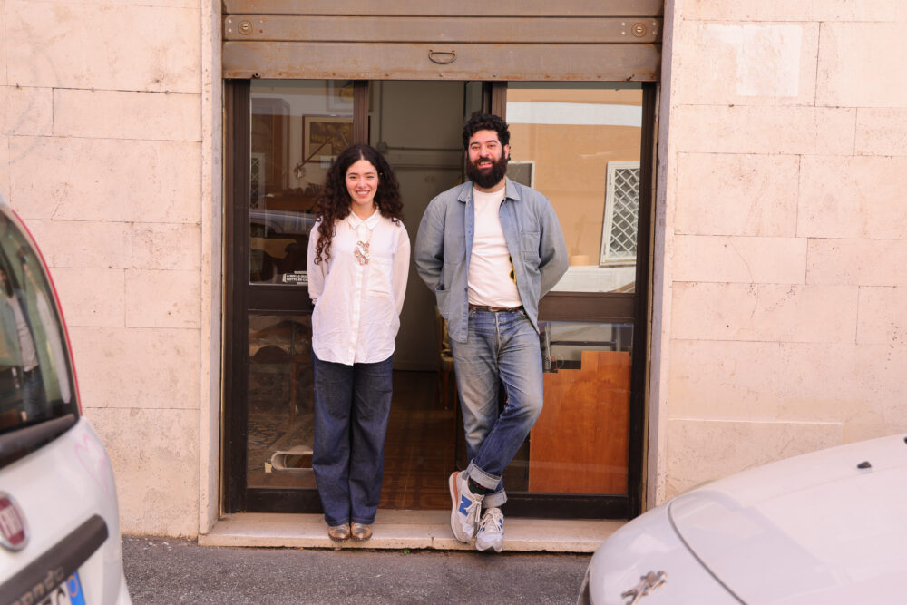 A woman in a white blouse and a man in a light blue jacket stand together at a doorway.