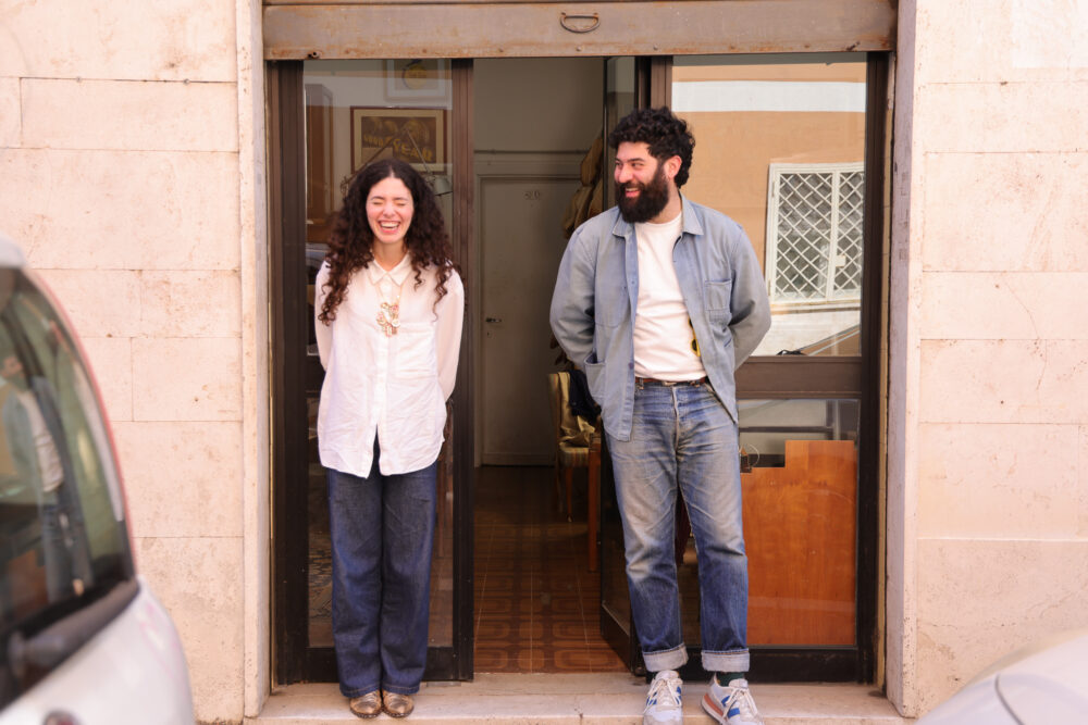 A woman in a white shirt and a man in a light blue jacket stand smiling at a doorway.