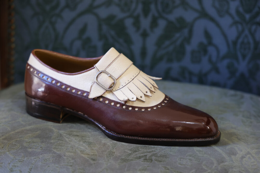 A polished brown leather shoe with a cream-colored strap and decorative fringe.