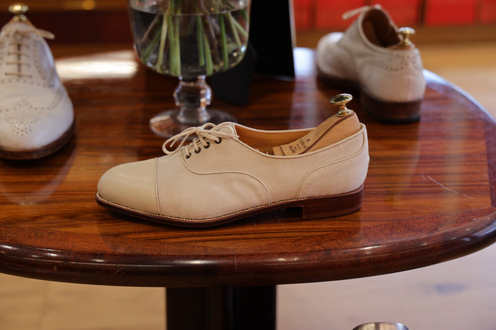 A cream-colored shoe with a leather toe and fabric body, featuring laces and a decorative heel.